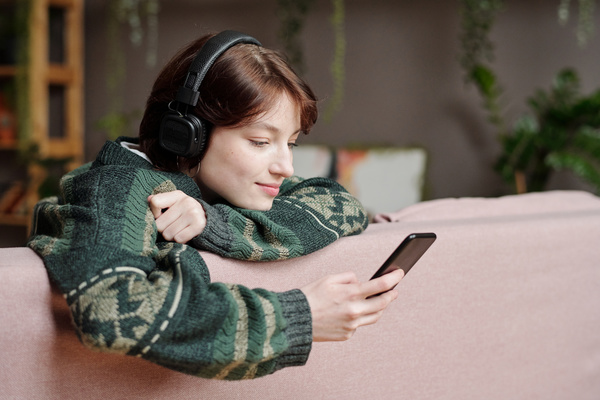A girl with short dark hair in black headphones wearing an ornamental sweater is watching a video on her phone sitting on the sofa and leaning on its back